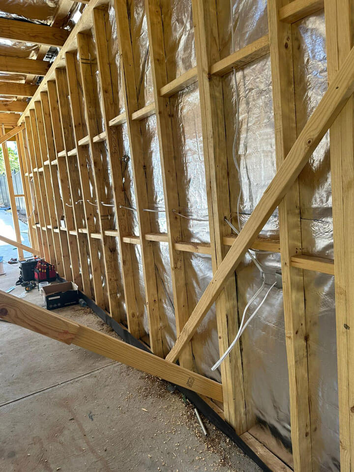 Partially constructed wooden wall frame with insulation installed, showing exposed timber studs and electrical wiring in progress.
